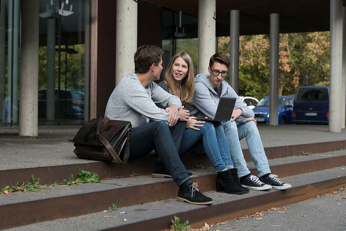 Drei Studenten sitzen gemeinsam auf einer Treppe und arbeiten an einem Laptop.